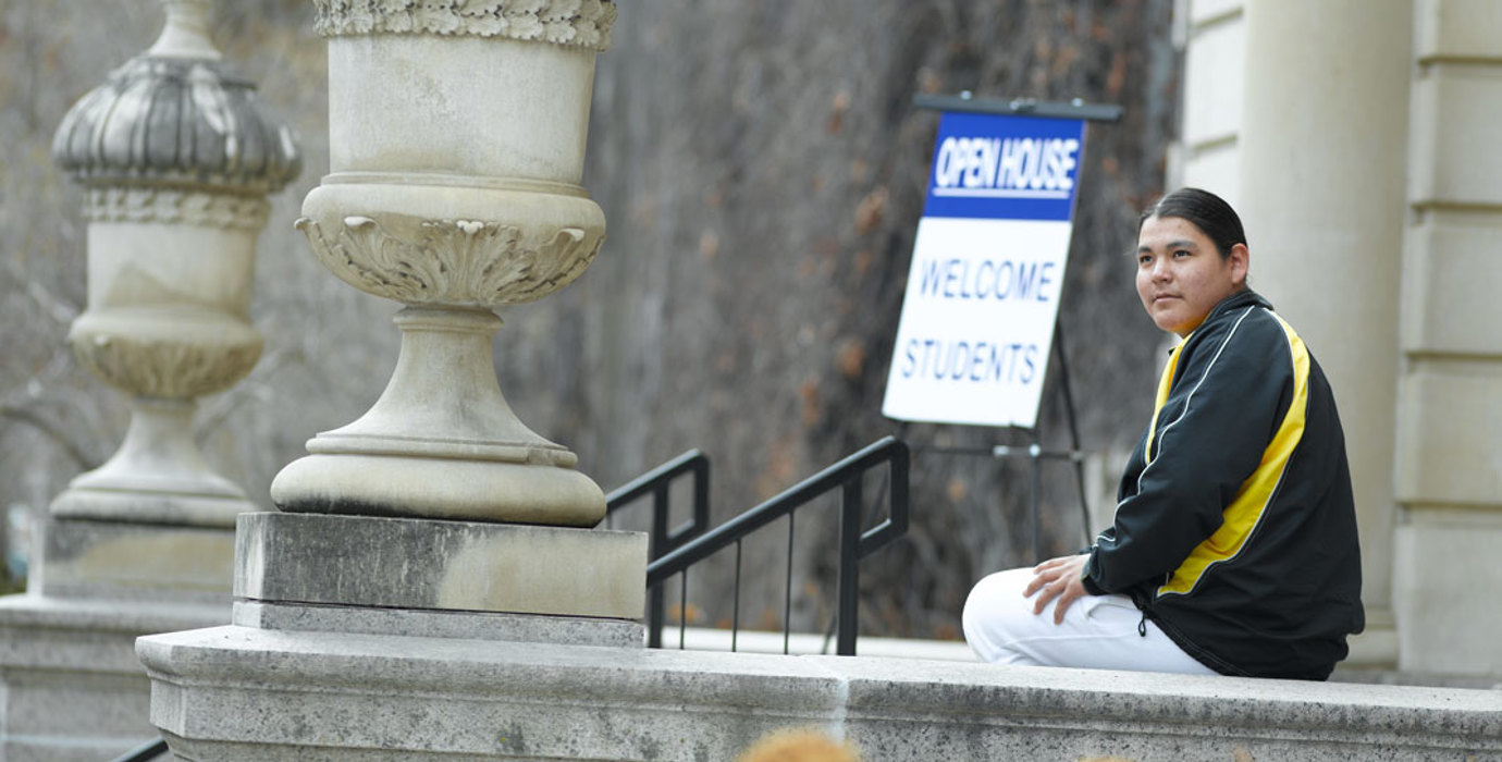 Student sitting outside a school building with an "Open House: Welcome Students" sign in the background