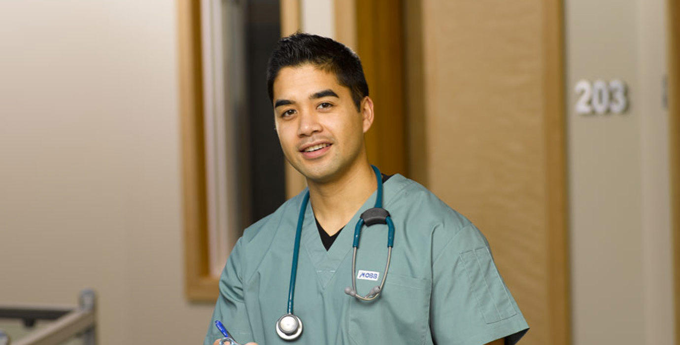 Nurse wearing scrubs with stethoscope around neck, carrying a clipboard stands in a hospital hallway