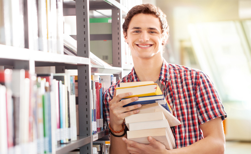 Youth holding a stack of books while leaning against a library shelf