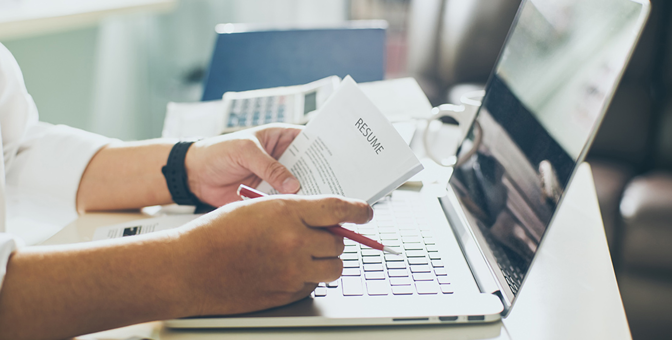 Hands holding a resume and pencil on top of an open laptop