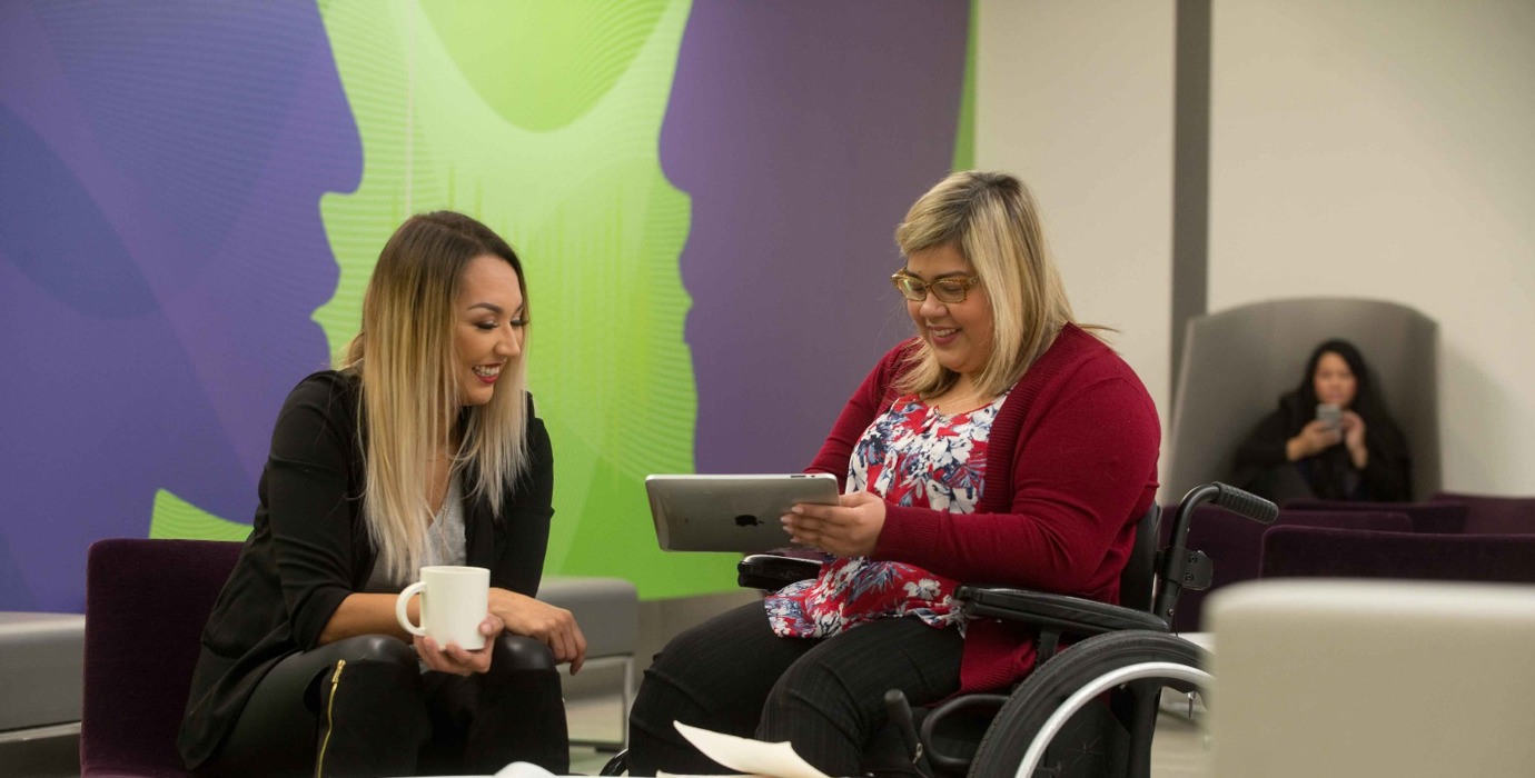Student holding a tablet while chatting with a friend in a campus common room