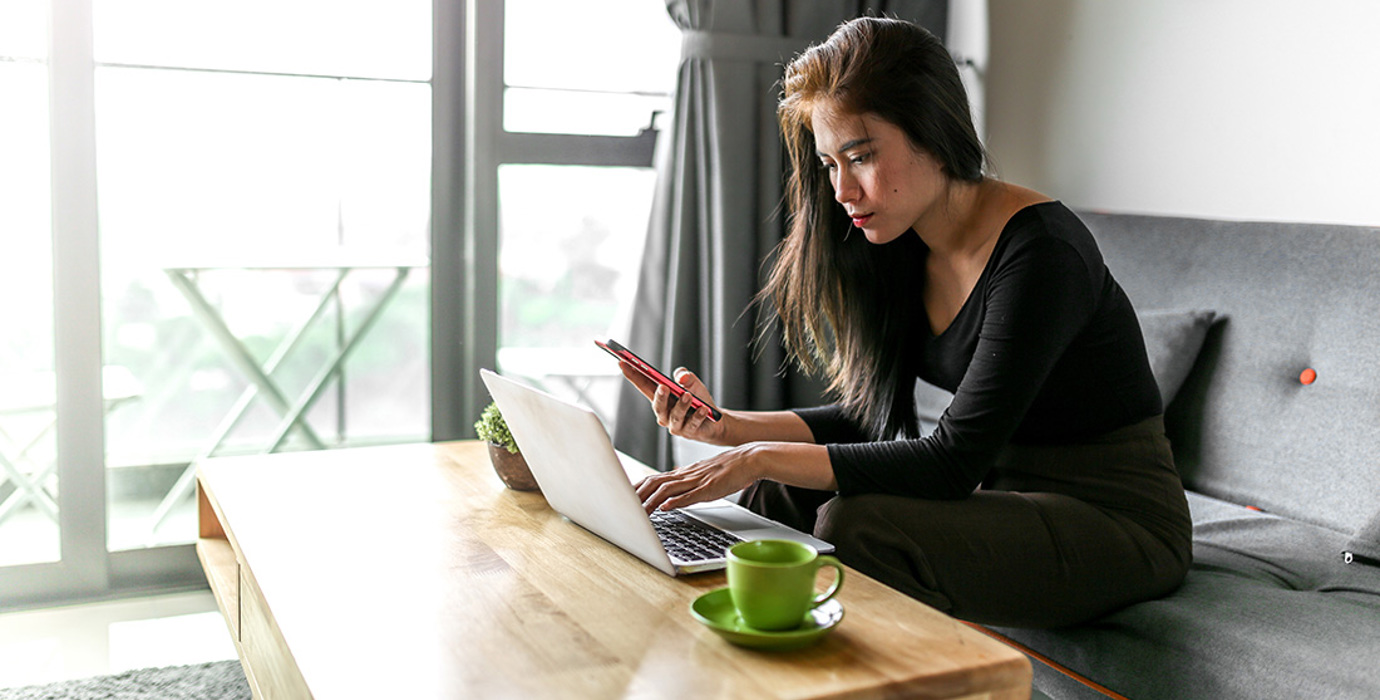 Person sitting on a couch in the living room looking at a laptop and smartphone