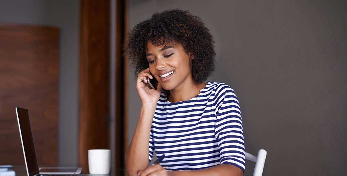 Person wearing a striped shirt, sitting at a table and talking on the phone while taking notes