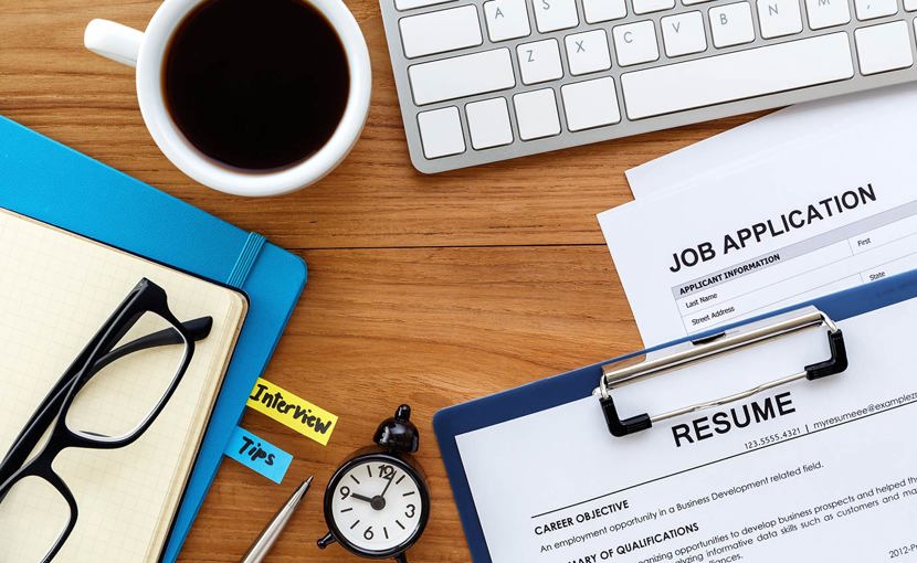 Resume, job application, notebooks, keyboard, coffee and a clock laid out on a desk