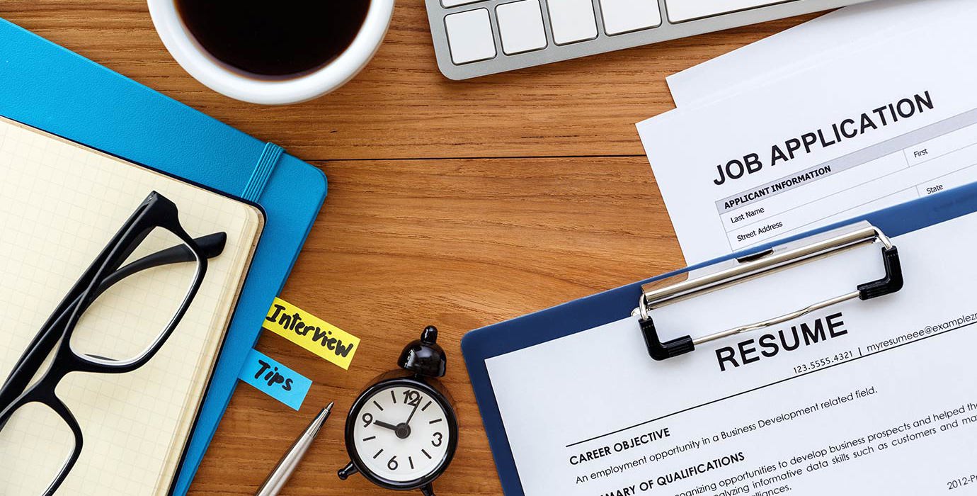 Resume, job application, notebooks, keyboard, coffee and a clock laid out on a desk