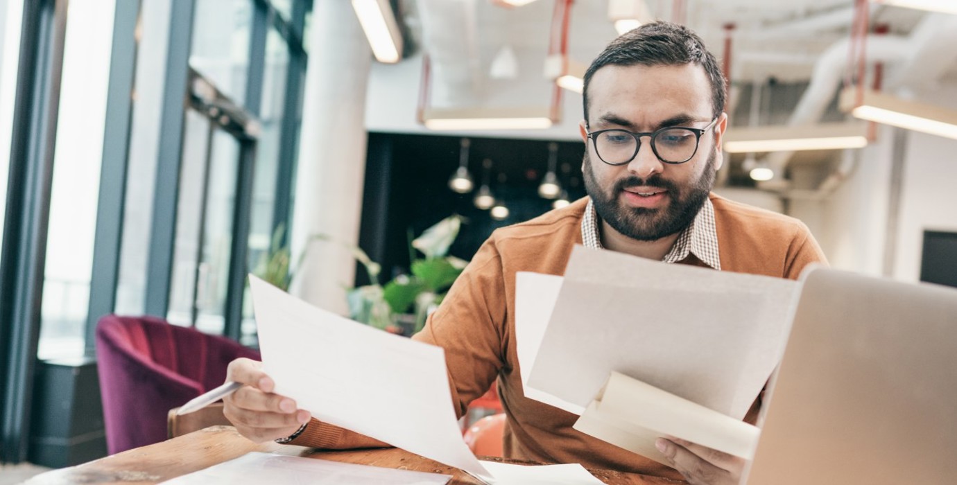 Person sitting at computer and looking through several pieces of paper.