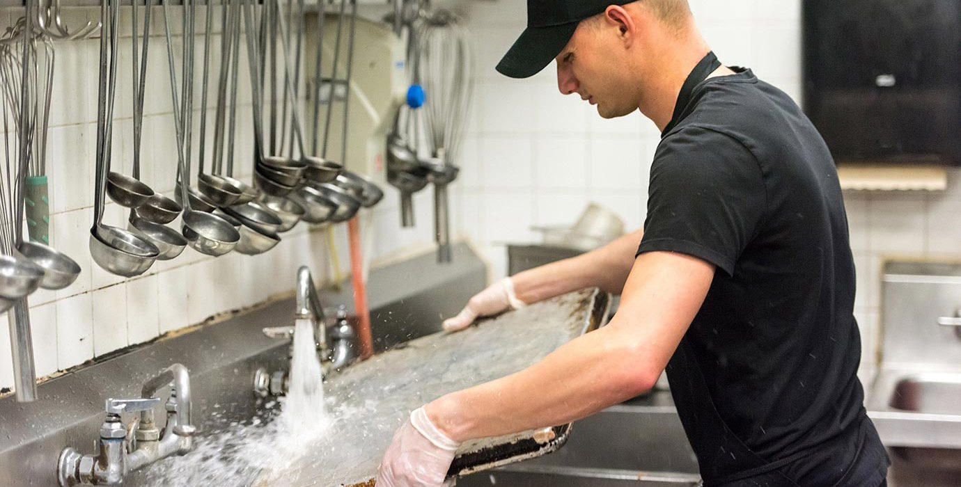 Man washing baking sheet in the sink of a commercial kitchen.