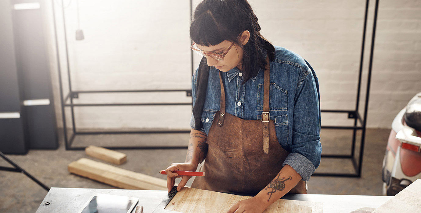 Woman doing woodworking while looking at a tablet in her workshop.