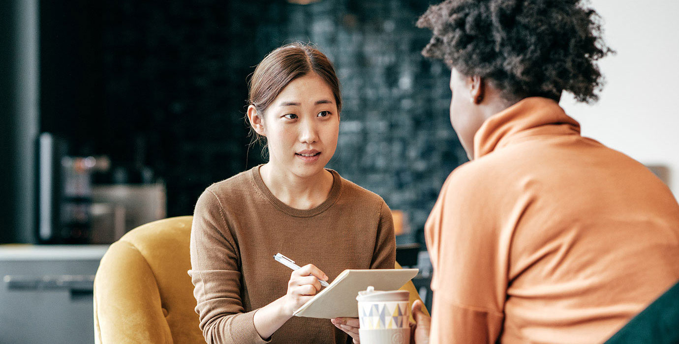 Woman interviewing another woman and taking notes as she speaks. 