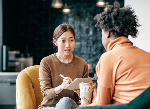 Woman interviewing another woman and taking notes as she speaks. 