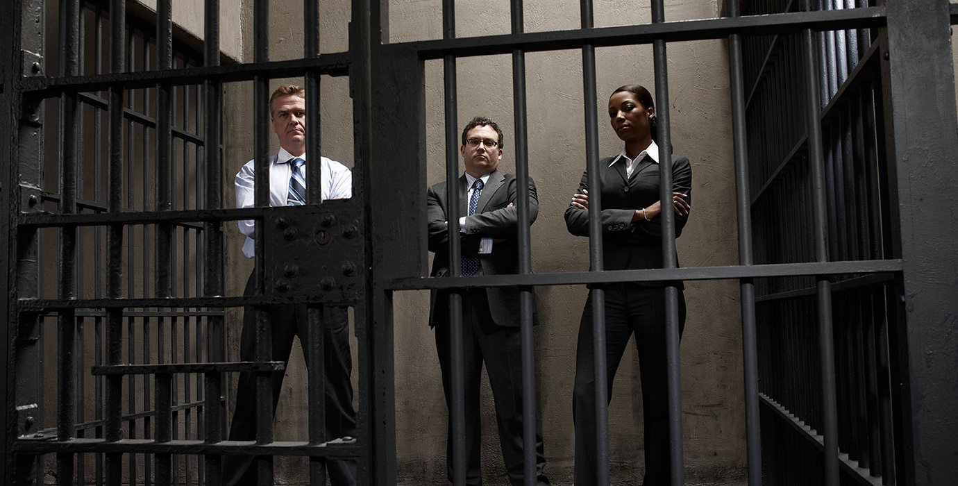 3 people wearing business suits with arms crossed standing inside a prison cell.