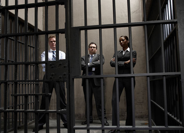 3 people wearing business suits with arms crossed standing inside a prison cell.