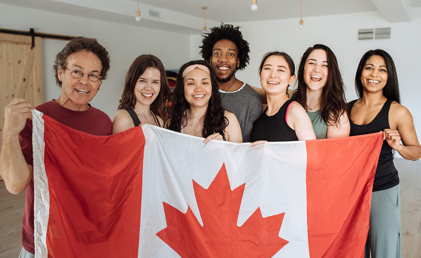 A happy group of multi-ethnic people stand behind a large Canada flag.