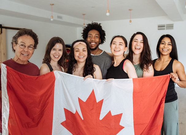 A happy group of multi-ethnic people stand behind a large Canada flag.