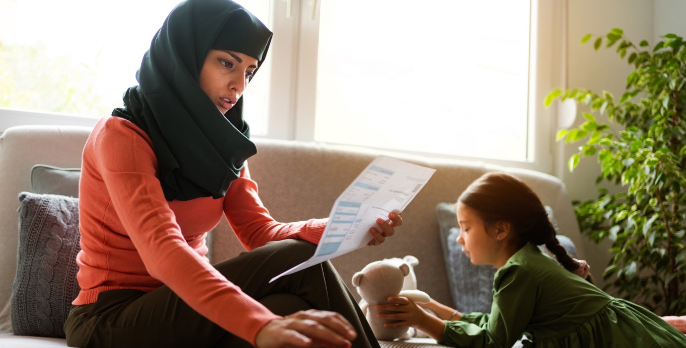 Mother paying bills while sitting on the couch with her young daughter, who is playing with a stuffed animal.