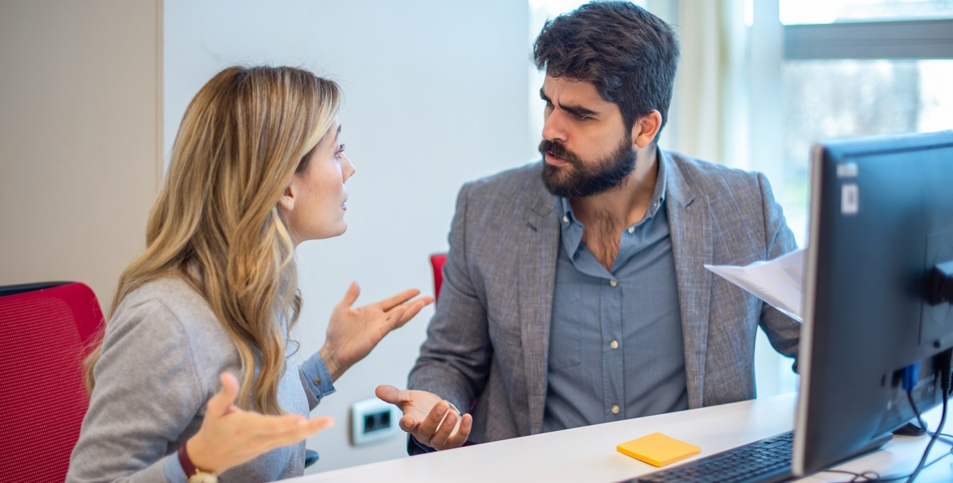 Two colleagues arguing while sitting at a desk at work. 