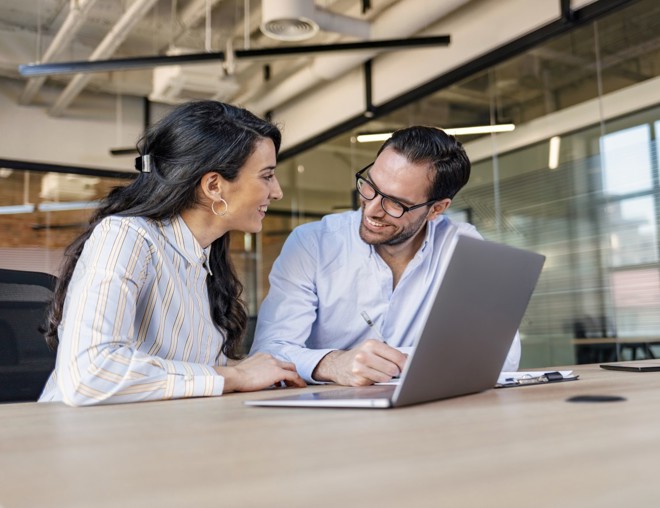 Two coworkers being friendly while sitting at a desk over a laptop. 