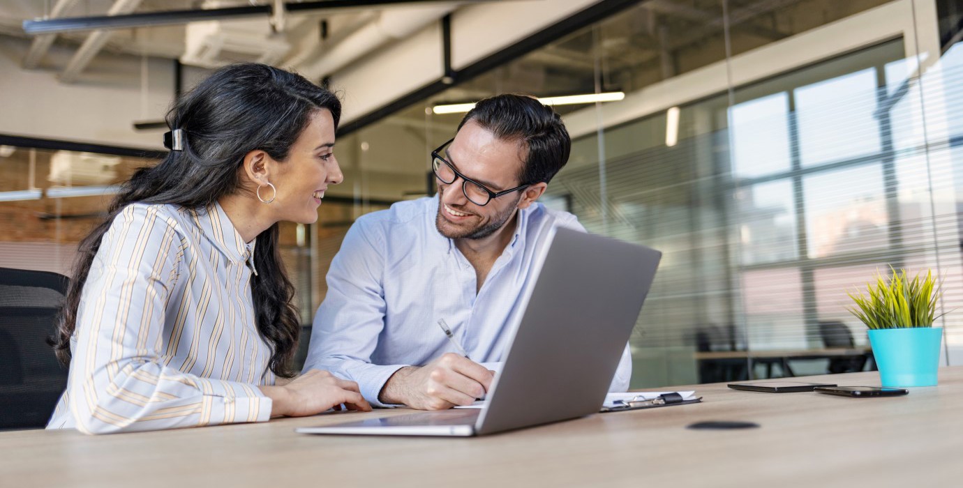 Two coworkers being friendly while sitting at a desk over a laptop. 